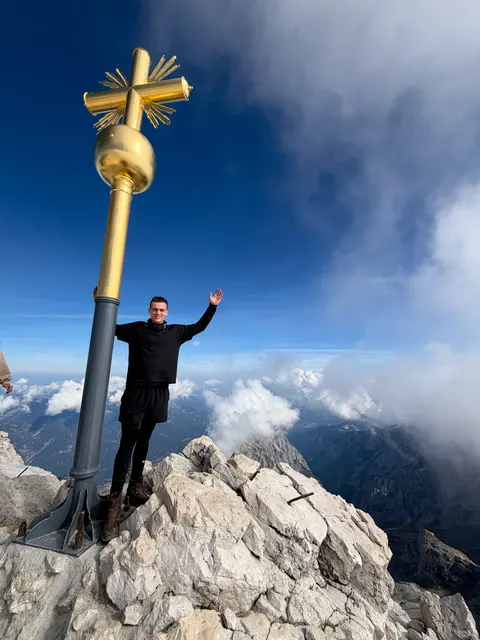 Ben mit erhobenen Armen am goldenen Gipfelkreuz der Zugspitze